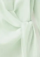Camisa cruzada de ramio en color crudo, con cuello camisero, escote en V y mangas largas con puños anchos abotonados. Cuenta con un diseño envolvente con detalle de lazo lateral.