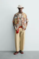 Camisa de corte relajado confeccionada en tejido ligero de algodón, con cuello bowling y manga corta. Presenta un estampado integral con motivos urbanos y tropicales en tonos cálidos.