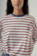 Camiseta de manga larga con diseño de rayas horizontales en color rojo sobre fondo blanco. Presenta cuello redondo en contraste azul marino y un pequeño bordado con la letra R en el pecho. Posee un corte holgado y relajado.