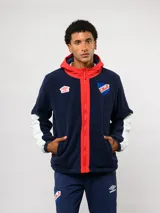 Campera de polar azul marino con capucha, cierre frontal y detalles en rojo. Presenta el logo de Umbro y el escudo del Club Nacional de Football en el pecho, con paneles blancos en las mangas.