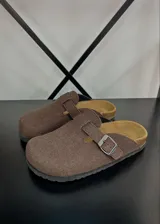 Zuecos de gamuza color marrón chocolate, con diseño de punta cerrada, plantilla anatómica y tira ajustable con hebilla metálica lateral.