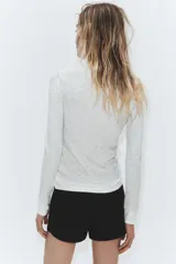 Camiseta de manga larga color gris melange, con cuello redondo y bajo recto. Presenta un detalle de tejido efecto doble a contraste en el cuello, puños y dobladillo inferior en color blanco.