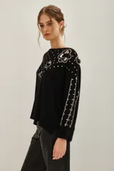 Sweater negro de punto con cuello redondo, manga larga y diseño holgado. Presenta aplicaciones de lentejuelas plateadas dispersas, bordes plateados en las mangas y apliques florales con lentejuelas en el hombro y frente.