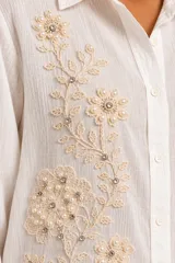 Camisa blanca de manga larga confeccionada en lino y viscosa, con cuello camisero, cierre frontal con botones y delicados bordados florales con apliques de perlas y pedrería en el frente.