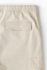 Pantalón cargo color beige de corte slim fit, confeccionado en tejido de algodón. Cuenta con cintura elástica ajustable con cordón, bolsillos al bies, bolsillos laterales y traseros con solapa y botones a presión ocultos, costuras y pinzas en las rodillas.