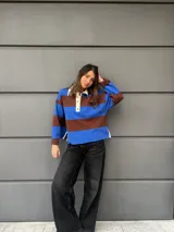 Buzo tipo rugby de algodón con diseño de rayas horizontales anchas en azul y marrón. Presenta cuello polo en color crema con cierre frontal de tres botones y mangas largas.
