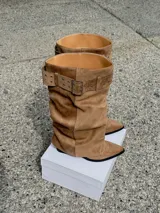 Botas altas de gamuza color beige, con diseño slouchy, punta fina y una tira ancha superior con hebilla metálica rectangular y ojalillos.