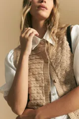 Chaleco tejido sin mangas color blanco roto, con cuello redondo y cartera frontal de botones. Presenta un punto calado con textura.