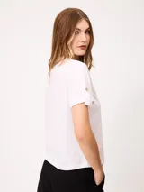 Remera blanca de manga corta con vuelta en los puños y detalle de botón dorado. Presenta cuello redondo y corte recto.