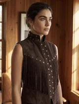 Chaleco de gamuza color marrón chocolate, sin mangas, con cuello mao, detalles de tachas metálicas en el cuello y la solapa frontal, y flecos delanteros de estilo western. Cuenta con cierre frontal de broches a presión.