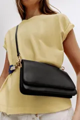 Bolso bandolera rectangular de color negro con solapa y cierre de botón imantado, herrajes metálicos dorados y compartimento interior con cremallera. Incluye una correa ancha ajustable y extraíble en combinación de colores azul y blanco.