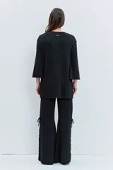 Cardigan negro de punto con acabado brillante, cuello redondo, cierre frontal de botones y detalle de cordones laterales entrelazados. Presenta mangas tres cuartos con diseño acampanado.