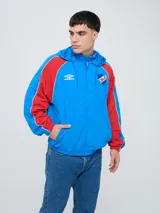 Campera deportiva Umbro del Club Nacional de Football, modelo Venti, para hombre. Diseño en color blanco con mangas azules y detalles de líneas rojas. Cuenta con capucha con cordón ajustable, cierre frontal y bolsillos. Presenta el logo de Umbro y el escudo del club bordados en el pecho.