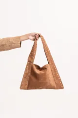 Cartera tipo tote de gamuza color tostado, con diseño triangular y correas integradas decoradas con tachas metálicas doradas a lo largo de los bordes.