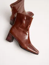 Botas de caña media color marrón, confeccionadas en cuero con acabado brillante. Presentan punta cuadrada, taco de madera y un distintivo bordado decorativo en color rojo que recorre el empeine hasta la punta.
