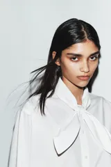 Camisa de popelín de algodón con cuello subido y detalle de aplicación tipo pañuelo anudado al cuello. Presenta manga larga con puños clásicos y cierre frontal mediante botones.