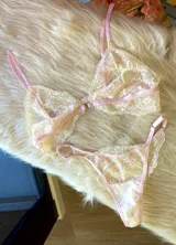 Conjunto de lencería compuesto por un bralette y una tanga, confeccionados en encaje bitono rosa y champagne con diseño floral. El bralette presenta cierre frontal con broche de gancho y breteles regulables. La tanga es de estilo colaless con laterales regulables.