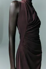 Vestido midi de color negro, con cuello subido y diseño sin mangas. Presenta un detalle de drapeado lateral en la parte delantera y cierre de cremallera oculta en la espalda.