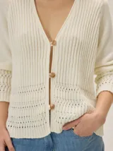 Cardigan de punto color blanco marfil, con escote en V, mangas largas con detalles de tejido calado y cierre frontal de botones con efecto madera.