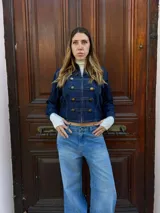 Chaqueta de denim azul oscuro con estilo militar, cuello alto, cierre frontal con cremallera y detalles de trabas decorativas con botones dorados.