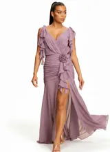 Vestido largo de fiesta color lila, con escote en V, mangas cortas con volados, detalle de frunces en la cintura y una flor decorativa lateral. Presenta una falda con tajo frontal y caída fluida.