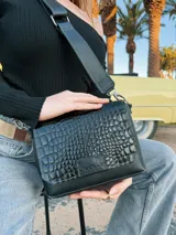 Bandolera negra de cuero con textura tipo cocodrilo en la solapa frontal, correa larga regulable y herrajes plateados.