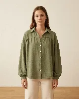 Camisa de calce amplio color verde oliva, con cuello camisero, hombros caídos y mangas largas con puños elásticos. Presenta detalle de frunce en el delantero y espalda, con cierre frontal de botones.