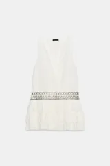 Vestido corto de gasa color blanco, con escote en V cruzado, sin mangas y diseño con volantes en el bajo. Presenta un detalle de tachas plateadas en la cintura.