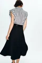 Vestido midi combinado con parte superior de camisa a rayas verticales blancas y negras, cuello solapa, manga sisa con frunces y botones dorados. La parte inferior es una falda midi negra de corte recto. Incluye un cinturón negro con textura de cocodrilo y hebilla dorada.