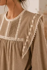 Blusa color tostado con cuello redondo, mangas largas abullonadas con puños elastizados, y detalles de puntillas y bordados en el frente y hombros.