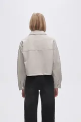 Chaqueta de eco-cuero en color beige, con diseño holgado, cuello tipo camisa y cierre frontal metálico. Presenta dos bolsillos frontales con solapa y mangas largas con puños rectos.