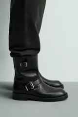 Bota de media caña en color negro, con diseño de horma cuadrada y suela con ligero tacón. Presenta dos cintas ajustables con hebillas metálicas, una en la parte superior de la caña y otra en la zona del empeine.