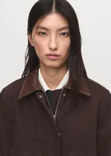 Chaqueta corta de algodón en color marrón oscuro, con cuello de efecto piel en tono contrastante. Presenta cierre frontal con cremallera y botones a presión, además de dos bolsillos frontales con solapa y cierre de botón.