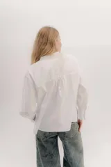 Camisa de algodón blanca con cuello a la base, cierre frontal con botones y mangas largas con puños delicados. Presenta volados verticales y detalles de cintas de terciopelo en contraste a lo largo del frente.