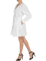 Vestido camisero corto color blanco, confeccionado en algodón orgánico. Presenta cuello en V, cierre frontal con botones, mangas largas abullonadas, cinturón de lazo ajustable en la cintura, bolsillos laterales y detalle de encaje calado en forma de escalera.