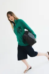 Cardigan de punto color marrón con cierre frontal, cuello alto y bolsillos aplicados. Presenta diseño tejido localizado con detalles en violeta y negro en el pecho.