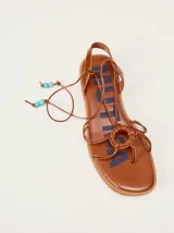 Sandalias planas de cuero color marrón, con diseño de punta cuadrada, tiras finas cruzadas y un detalle trenzado circular en el empeine. Cuentan con tiras para atar al tobillo decoradas con cuentas turquesas en las puntas.