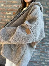 Campera tipo hoodie de corderito sintético en color gris, con cierre frontal, capucha y bolsillos laterales. Presenta un parche de cuero con el logo de la marca en la manga.