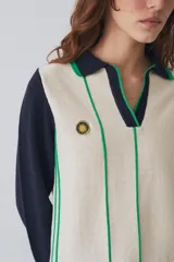 Remera tejida de punto estilo polo con mangas largas en color crema y cuerpo en color bordó con rayas verticales amarillas. Presenta cuello tipo polo, aberturas laterales y un pequeño parche circular con diseño de sol en el pecho.