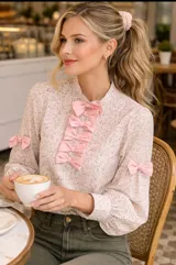 Blusa de cuello alto con estampado floral en tonos rosa y verde oliva. Presenta una hilera central de lazos decorativos en color rosa sólido, mangas largas abullonadas con puños ajustados y detalles de lazos adicionales en los antebrazos.