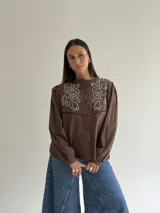 Camisa color marrón chocolate con bordado artesanal de flores y hojas en hilo crudo. Presenta cuello con volado, mangas largas con puños abotonados y cierre frontal con botones.