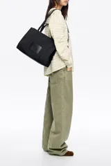 Bolso shopper de nylon negro, estructura blanda, con doble asa de mano y cierre principal de cremallera. Presenta una etiqueta logotipada en el frente y herrajes metálicos lacados.