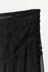 Falda de tiro bajo color negro, confeccionada en hilatura de viscosa. Presenta detalles de bordados y aplicación de puntilla a tono en la parte superior. El bajo tiene un acabado asimétrico con volados de gasa y cuenta con forro interior. El cierre es lateral con botones forrados.