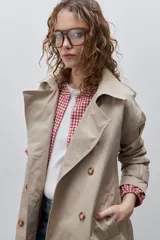Trench clásico color marrón, de silueta relajada, confeccionado en tejido liviano, con cuello de solapa, doble abotonadura frontal y mangas largas con presillas regulables en los puños.