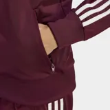 Campera deportiva Adidas modelo Firebird de corte holgado, color bordó con las icónicas tres rayas blancas en las mangas y logo del trébol bordado en el pecho. Cuenta con cierre frontal completo, cuello alto y puños acanalados.