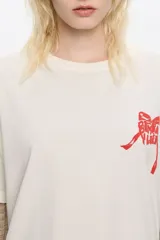 Camiseta de algodón color blanco hueso, de corte boxy y holgado, con escote redondo y mangas cortas. Presenta un pequeño logo en relieve de color rojo en el área del pecho.