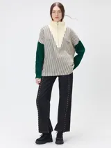 Pullover de tejido acanalado grueso con cuello alto y cierre frontal parcial. Presenta un diseño de bloques de color, con cuerpo en tono beige y mangas en contraste con rayas verticales en tonos rojizos. Incluye un detalle bordado en el pecho.