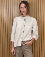 Camisa de lino color beige, con cuello alto mao con volados, mangas tres cuartos abullonadas y cierre frontal de botones oscuros.