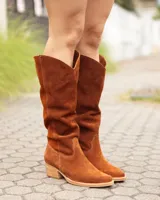 Bota estilo texana de caña alta, confeccionada en cuero gamuzado color marrón óxido. Presenta un diseño de corte en V en la parte superior de la caña, costuras decorativas y un taco bajo de madera.