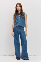 Chaleco de denim azul sin mangas, con cierre frontal de botones metálicos, bolsillos con solapa en el pecho y cuello camisero marrón de pana. Presenta bordados pequeños dispersos en color marrón.